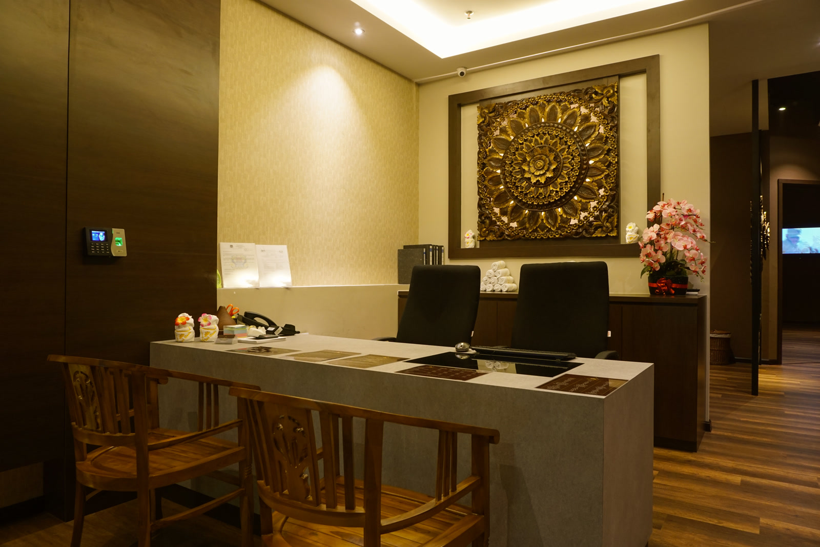 The Thai Oasis reception at Elit Avenue Penang: warm teak chairs, a quiet empty desk, and a carved gold Thai relief glowing on the wall behind it